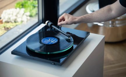Giradischi nero con disco nero ed etichetta blu, braccio di lettura regolato da una mano, posizionato su un mobile bianco.