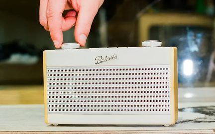 Bärbar retro-radio, krämfärgad rektangulär låda med ljus trä på sidorna och ett stort perforerat metallgaller på framsidan, två cirkulära knappar på ovansidan, framifrån sedd