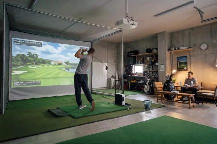 Indoor golf simulator with course screen, player mid-swing, and green putting mat
