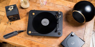 Mat sort Elipson pladespiller på træbord, omgivet af en sort højttaler, en vinylplade og hi-fi-tilbehør