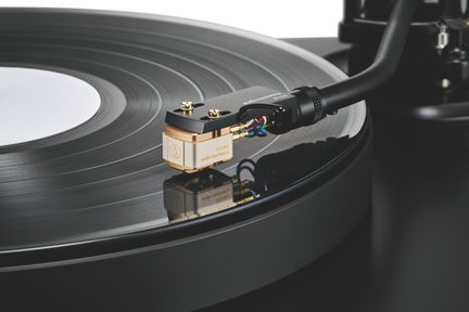 Primer plano de un disco de vinilo negro con surcos sobre un giradiscos, cápsula fonográfica de metal dorado con cables multicolores y brazo negro