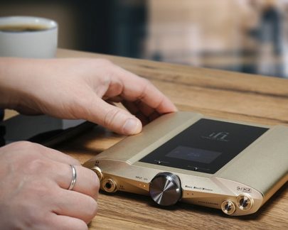 Zlatý audio přístroj s černou obrazovkou, dvěma kulatými tlačítky a jack konektory položený na dřevěném stole s rukou na něm