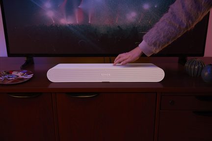 Fehér műanyag soundbar lekerekített élekkel, elölnézet, középen Sonos logó, fa bútoron, kéz a tetején