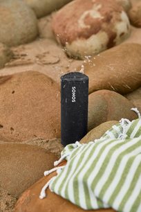 Sonos Roam Shadow Black Bluetooth-højttaler placeret på sand, ved siden af et grønt og hvidt stribet stykke stof og en menneskelig arm