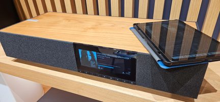 Tredjehånds skrå foroven-visning af en rektangulær højttaler med lyst træ på toppen, front i antracitgråt stof med et centralt farvedisplay og en række berøringsfølsomme knapper, en sort tablet placeret ovenpå
