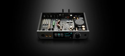 Carcasă audio din metal negru și aluminiu argintiu, vedere frontală ușor de sus, capacul îndepărtat dezvăluind placa electronică cu șiruri de condensatori aurii și verzi, radiator circular mare și panou frontal cu ecran color, selector mare negru și două mufe pentru căști aurii