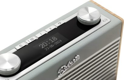 Blå ankafärgad bordradio Roberts Rambler Uno, rektangulär låda klädd i konstläder med ljus träkant, front med perforerat galler, svart digital display omgiven av vita knappar och två aluminiumreglage, trekvarts vy från ovan