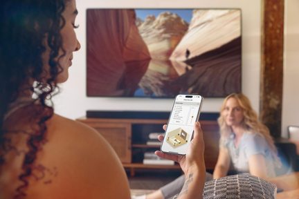 Vedere de aproape a unei mâini cu smartphone argintiu cu margini metalice, ecran de aplicație, TV montat pe perete, mobilier din lemn