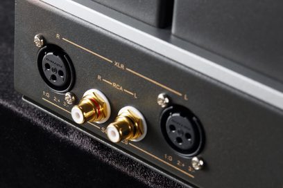 Schuine close-up van het grijze metalen achterpaneel van de Cayin HA-2A, een high-fidelity hoofdtelefoonvoorversterker, uitgerust met twee zwarte XLR-aansluitingen bevestigd met chromen schroeven en twee vergulde RCA-connectoren met witte ringen.