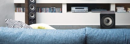 Garso sistema su garsiakalbiais, stiprintuvu, CD grotuvu TV spintelėje su lentynomis, kuriose sudėti CD ir knygos, prieš ją mėlyna sofa