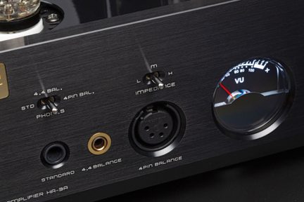 Primo piano frontale su un pannello anteriore in metallo spazzolato nero di amplificatore audio con prese cuffie standard e bilanciate, connettore XLR a 4 poli, due interruttori a levetta argentati e un VU-meter circolare retroilluminato