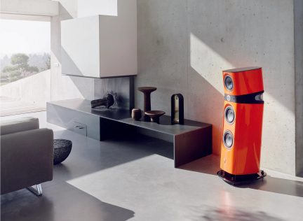 Leuchtend orangefarbener Lautsprecher mit drei runden Lautsprechern, rechts auf grauem Boden positioniert, Betonwand im Hintergrund