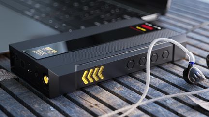 Caja de audio portátil negra de aluminio, vista en tres cuartos, con flechas amarillas retroiluminadas en el frontal, panel superior estriado e inserto de vidrio negro, cuatro botones hexagonales laterales
