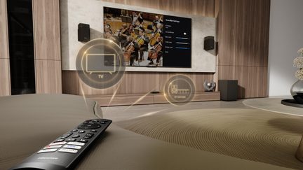 Udsigt fra sofaen, sort plastikfjernbetjening med farvede knapper i forgrunden, stor vægmonteret fladskærm indbygget i et lyst træpanel med to væghøjttalere og en subwoofer, gennemsigtige cirkulære ikon-haloer svævende foran