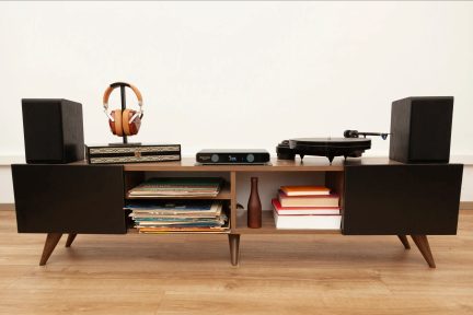 Vedere frontală a unui dulap jos din lemn maro, cu uși negre și picioare subțiri, care are în compartimentele deschise discuri de vinil și cărți, în timp ce pe blat se află două boxe negre, o pereche de căști maro pe suport, o carcasă electronică neagră și un pickup negru.