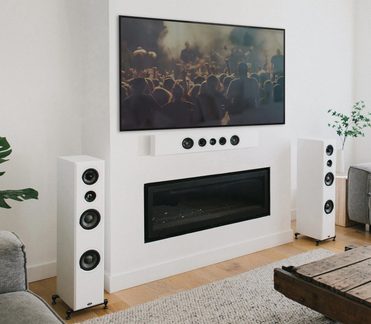 Vista frontal de uma sala, TV acima de uma lareira preta, colunas de som brancas, piso de madeira