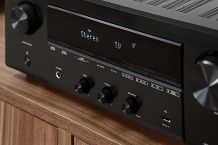 Sort AV-forstærker i metal, set i trekvart forfra, display “Stereo TV”, store drejeknapper, hovedtelefonstik og USB-port