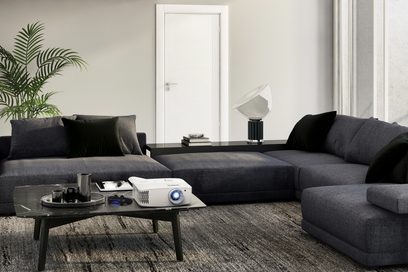 Proyector Optoma UHD35 blanco y negro colocado sobre una mesa baja negra en una sala de estar moderna con un sofá de esquina gris oscuro
