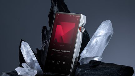 Lettore audio portatile argentato in metallo con spigoli angolati, schermo touch nero attivo, rotella laterale zigrinata con bordo rosso, visto da tre quarti davanti, posizionato tra cristalli traslucidi
