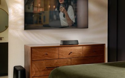 Træmøbel til TV med skuffer, sort Polk Audio MagniFi Mini AX soundbar placeret ovenpå, TV-skærm der viser en kvinde der drikker kaffe