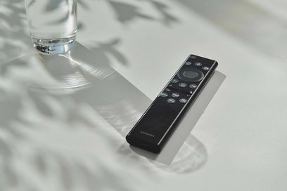 Černá dálkový ovladač Samsung položený na odrazivém povrchu vedle průhledné sklenice vody z boku