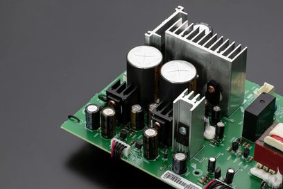 Placa de circuito impresso verde com componentes eletrônicos, dissipadores de alumínio e dois grandes condensadores cilíndricos prateados, vista superior