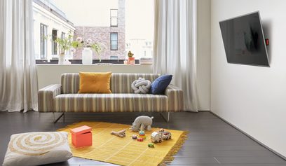 Moderne stue med stribet sofa, farvede puder, gult tæppe, legetøj på gulvet, hvide gardiner og vægmonteret tv