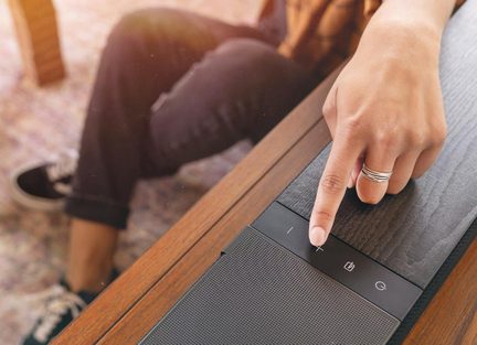 Bară de sunet neagră pe un mobilier din lemn, mână feminină apăsând pe butonul tactil de volum plus