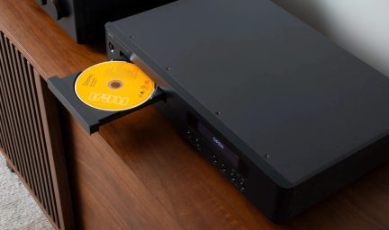 Czarny odtwarzacz CD NAD widziany pod kątem trzech czwartych, z otwartą tacką i żółtą płytą, na brązowym meblu