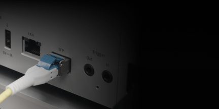 Boîtier électronique gris métal, vue de face en gros plan, ports USB et RJ45 noirs, logement SFP argenté abritant un connecteur fibre blanc à languette bleue, deux prises jack Trigger noires