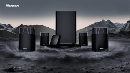 Sort hjemmebiograf-højttalersæt i tekstureret plast med metalgitre, bestående af en central, rektangulær subwoofer, fire kubeformede satellitter med topventilation og en lille, flad boks i forgrunden, vist forfra
