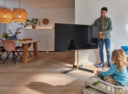 Wspornik ścienny Vogel's TVS 3695 w kolorze dębowym na płaski telewizor, z mężczyzną i dziewczynką w nowoczesnym, jasnym salonie