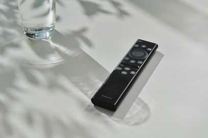 Sort Samsung fjernbetjening med knapper på hvid baggrund, set forfra, liggende ved siden af et glas vand på en reflekterende overflade