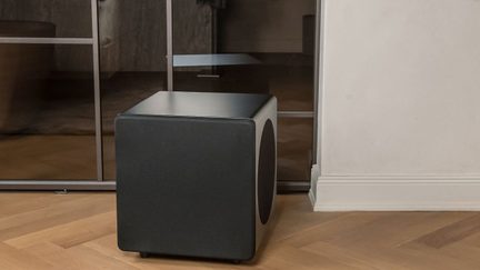 Minimalistinio dizaino juodas kubo formos žemųjų dažnių garsiakalbis ant grindų priešais vidinę vitrininę sieną