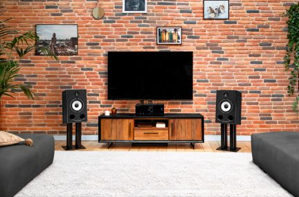 Par de altavoces Triangle negros sobre soportes, con conos blancos, a ambos lados de un televisor en un salón