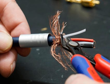 Primo piano obliquo su un tronchese in acciaio con manici rossi e blu che taglia lo schermo in fili di rame di un cavo audio guainato blu dotato di un manicotto metallico argentato, con conduttori interni rossi, neri e bianchi visibili.