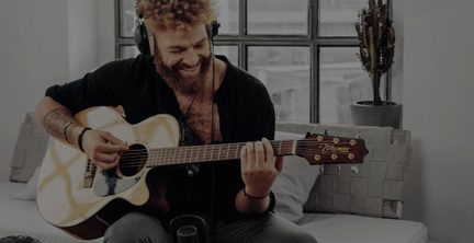 Kestirme panelli, cilalı açık renk ahşap akustik gitar çalan, siyah kulaklık takan ve gülümseyen oturmuş bir adamın üç çeyrek önden görünümü