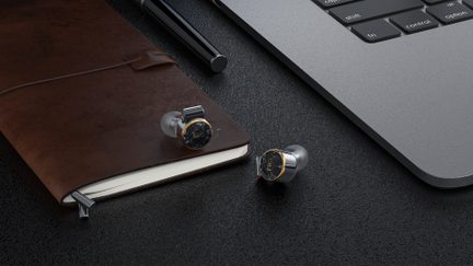 Pair of silver metal in-ear headphones with translucent silicone tips and a black marbled front surrounded by a gold ring featuring the “FiiO” logo, top view on a textured black surface
