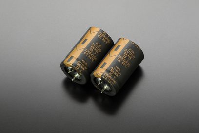 Dva zlaté válcové elektrolytické kondenzátory, viděné z boku, označené "FOR AUDIO 2200μF 63V", položené na šedém povrchu