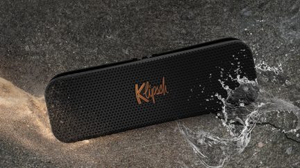 Altoparlante Bluetooth portatile Klipsch nero con griglia perforata, appoggiato sulla sabbia bagnata con schizzi d’acqua