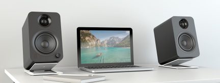 Graues Aluminium-Laptop mit geöffnetem Bildschirm, der eine Wasserlandschaft zeigt, auf einem weißen Schreibtisch zwischen zwei mattschwarzen, würfelförmigen Lautsprechern auf geneigten Ständern, Frontalansicht