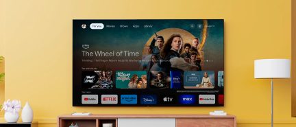 Fladskærms-tv monteret på væggen, der viser Google TV-interface, gul væg, lavt træmøbel, hvid lampe, hvid vase med blomster