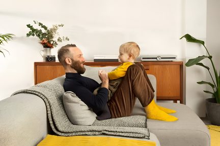 Grå stofsofa med plaid, skægget far og blondt barn siddende i lys, moderne stue