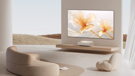 Nowoczesny salon z beżową, zakrzywioną kanapą, dopasowanym fotelem, ekranem ściennym wyświetlającym kwiaty, meblem wiszącym, projektorem, białymi zasłonami