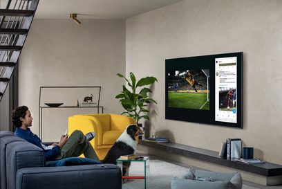 Mand sidder på en grå sofa i en moderne stue og ser på en stor vægmonteret tv, der viser en fodboldscene