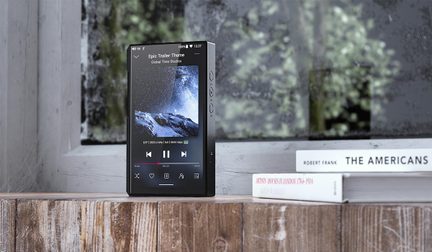 Rektangulær digital afspiller i mat sort aluminium, tændt touchskærm med lydafspiller vist, højre side med tre knapper, stående oprejst på en træoverflade, set fra trekvart forfra