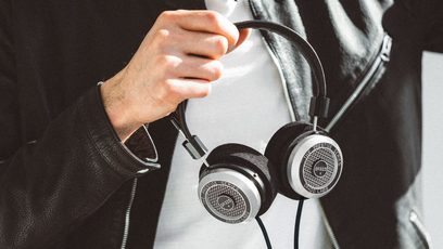 Lähikuva Grado SR325x -kuulokkeista sivulta katsottuna, harmaan neuleen päällä oleva käsi pitelee niitä, korvakupujen metalliritilät näkyvissä