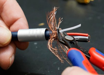 Tæt sidebillede af en blå lydkabel med cylindrisk sølvmetalhus, kobberskærmningsfletninger udfoldet omkring interne ledninger i rød, sort og hvid, holdt fast af en sort kabelsaks med blå og røde greb