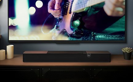 Černý obdélníkový soundbar Klipsch na dřevěném TV stolku, pod velkou plochou obrazovkou se záběrem zblízka na hráče na elektrickou kytaru