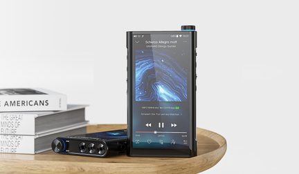 Tredjehåndsvisning af en sort, bærbar lydafspiller i metal og glas med stor touchskærm, der viser en musikgrænseflade, rillet drejehjul med blå kant øverst til højre og fysiske sideknapper, et andet lignende apparat ligger ved siden af på et lyst træfad